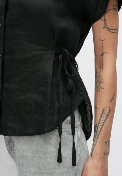 Μαύρη λινή μπλούζα με κοντά μανίκια, πλαϊνά κορδόνια για ρύθμιση και υφασμάτινη υφή, συνδυασμένη με ανοιχτό γκρι τζιν.