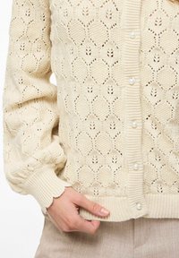 Gros plan sur une main tenant l'ourlet d'un cardigan en maille couleur crème avec des boutons en perle et des motifs de diamants texturés.