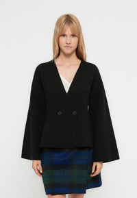 Black double-breasted jacket with wide sleeves, made of a textured fabric. Worn over a white top, paired with a blue and green plaid skirt.