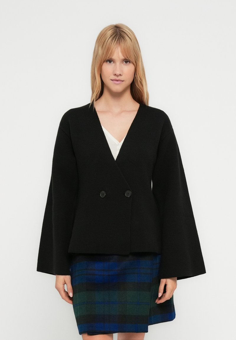 Black double-breasted jacket with wide sleeves, made of a textured fabric. Worn over a white top, paired with a blue and green plaid skirt.