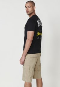 Camiseta negra de manga corta con estampado gráfico en la parte posterior, combinada con pantalones cortos de carga beige que cuentan con múltiples bolsillos y detalles de costura.