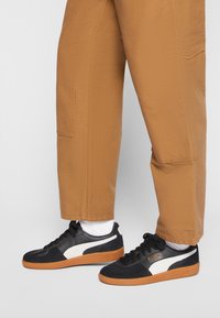 Sneakers bianco e nero con tomaia in pelle testurizzata, dotate di suola in gomma arancione. Abbinati a pantaloni larghi color sabbia e calze bianche.