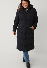 Lange zwarte pufferjas met een capuchon, voorzien van gestikte secties, ritsluiting en zijzakken. Draagt over blauwe spijkerbroek en enkellaarsjes.