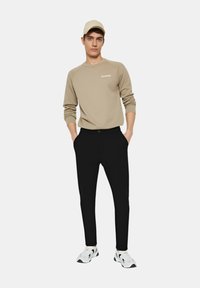 Felpa beige con logo, pantaloni neri e sneakers bianche con accenti neri. Uomo in piedi con le mani in tasca, indossa un cappello beige.