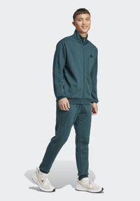 Survêtement vert foncé en tissu doux ; veste zippée avec col montant et logo ; pantalon slim ; associé à des chaussures de sport blanches.
