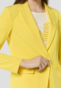 Blazer jaune avec col à revers crantés, fermeture à un bouton et deux poches avant, en tissu lisse. Superposé à un haut clair.
