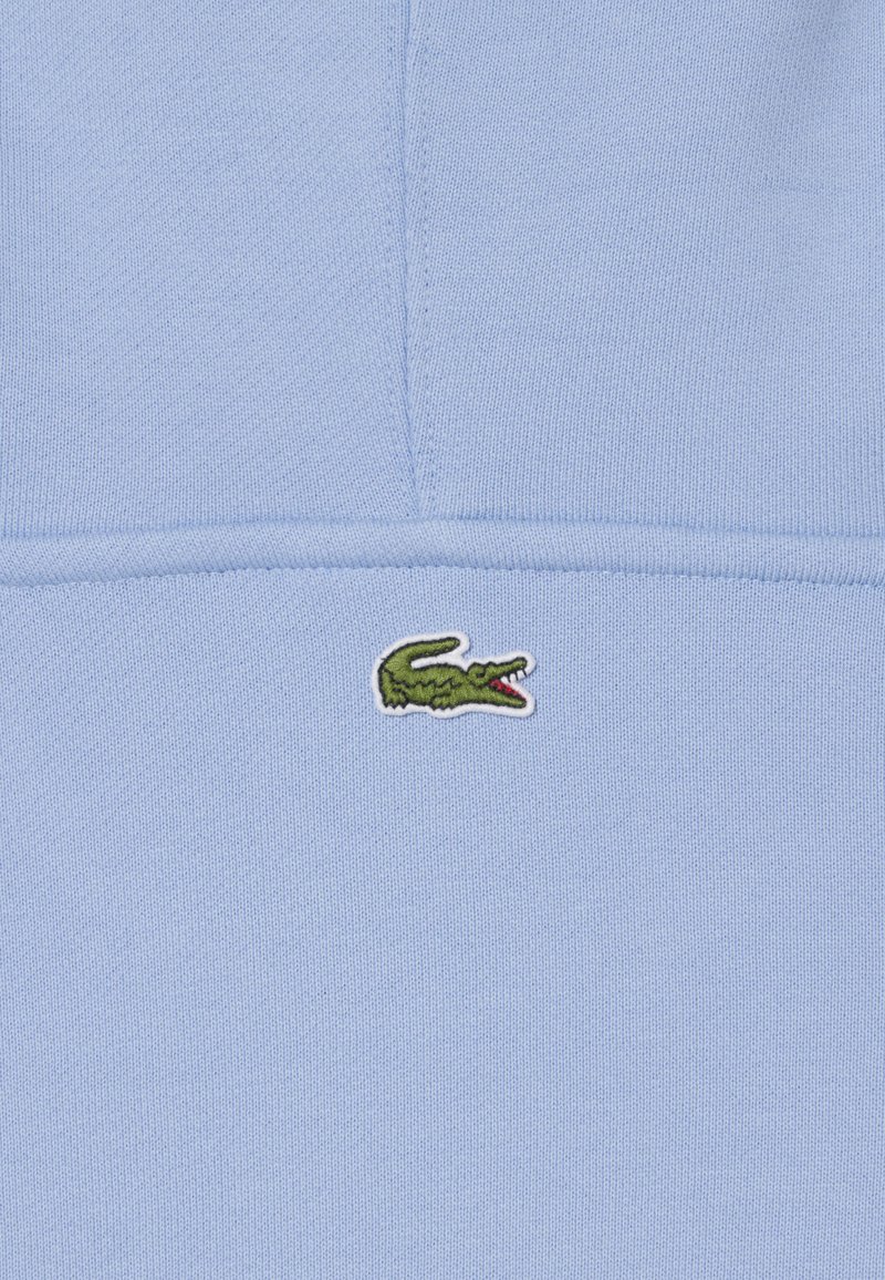 Tessuto azzurro chiaro con un piccolo logo di coccodrillo verde ricamato, centrato vicino a una cucitura.