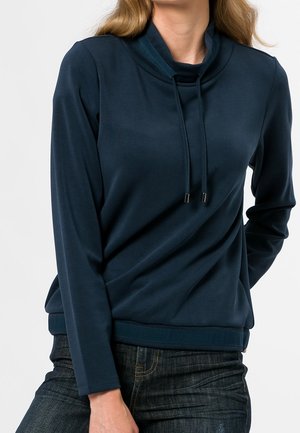 Kobieta w granatowej, długiej bluzie z golfem i sznurkami, w połączeniu z ciemnymi jeansami, na białym tle.