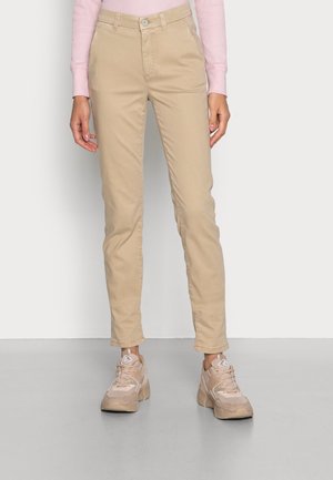 Persoon die beige slim-fit chino's, een lichtroze top met lange mouwen en beige chunky sneakers draagt, staand op een grijze vloer.