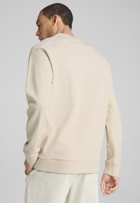 Sudadera beige con ribetes en el dobladillo y en los puños, textura suave, mangas largas y diseño de cuello redondo. Vista trasera, combinada con pantalones blancos.