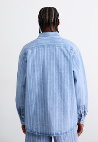Lyseblå denimskjorte med lodrette hvide striber, lange ærmer, knapmanchetter og klassisk krave, set bagfra.
