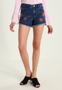 Džínové šortky s roztrhaným lemem, zdobené květinovou výšivkou v červené a zelené barvě. Skombinované s černými nazouváky s otevřenou špičkou a růžovým topem s dlouhým rukávem.