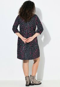Schwarzes Kleid mit pinkem und grauem Leopardenmuster, geraffter Taille, 3/4-Ärmeln und knielangem Saum; kombiniert mit metallischen Knöchel-Stiefeln.