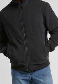 Persona in giacca di pile nera con le mani in tasca, che indossa jeans di denim blu, contro uno sfondo semplice.