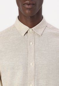Camisa de botón color beige claro con una textura suave, cuello con solapa y un sutil detalle de costura. Presenta botones color crema a lo largo del frente.