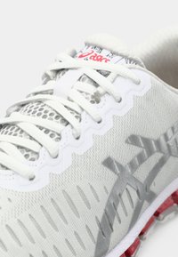 Chaussure de running ASICS blanche en gros plan montrant une tige en mesh, des lacets blancs, un logo gris et des accents rouges sur la semelle.