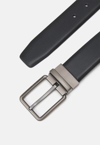 Ceinture noire texturée avec une boucle rectangulaire en argent mat, comportant une gravure discrète de la marque. Comprend plusieurs trous d'ajustement.