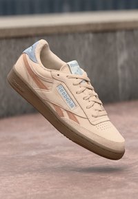 Beige Wildledersneaker mit hellblauen Akzenten und braunen Streifen, ausgestattet mit einer strukturierten Gummisohle und einem Logo an der Seite.