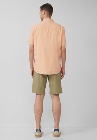 Korte mouwen, licht perzikkleurige overhemdd, olijfgroene shorts, beige schoenen met marineblauwe accenten, staand op een neutrale achtergrond.