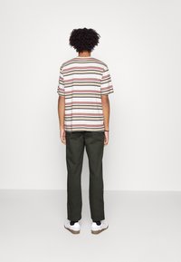 T-shirt rayé en crème, bordeaux et noir ; coupe décontractée avec des manches courtes. Un pantalon vert olive et des chaussures blanches complètent la tenue.