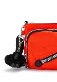 Borsa in tessuto arancione con hardware nero e tasca con zip. È attaccato un portachiavi a forma di gorilla peloso nero. Tessuto testurizzato con dettagli.