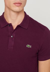 Burgundská polo košile vyrobená z bavlny, s klasickým límcem, zapínáním na dva knoflíky a zeleným krokodýlím logem na prsou.