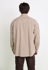Man stands facing away wearing a beige and white vertical striped long-sleeve shirt and dark brown cargo pants against a plain background.