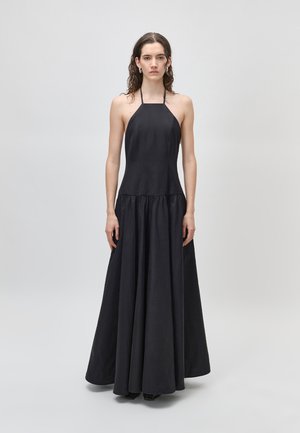 Hosszú, göndör hajú nő ujjatlan, fekete halternyakú ruhában, egyszínű világos háttér előtt áll.