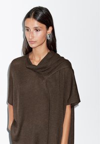 Kobieta z długimi ciemnymi włosami nosi duże, srebrne, owalne kolczyki oraz brązowy sweter dzianinowy z opadającym dekoltem na białym tle.