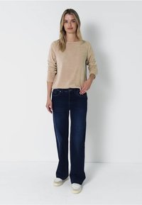 Beige Langarmoberteil aus weichem Stoff, kombiniert mit dunkelblauen, ausgestellt geschnittenen Jeans. Das Modell steht vor einem schlichten weißen Hintergrund.