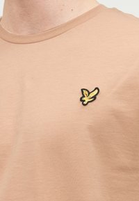 Φωτεινή μπεζ μπλούζα από απαλό βαμβάκι, με κεντητό λογότυπο πουλιού σε μαύρο και κίτρινο στο στήθος, ριμπ λαιμόκοψη.