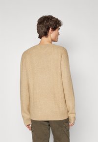 Pull en tricot beige avec un motif de câble texturé, col rond et manches longues, présentant des poignets côtelés et une coupe décontractée.