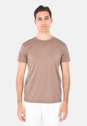 Giovane uomo con capelli corti e scuri, che indossa una maglietta marrone a maniche corte e pantaloni bianchi, in piedi davanti a uno sfondo chiaro.