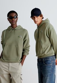 Dos modelos llevando sudaderas con capucha. El primer modelo tiene una sudadera verde oliva con un bolsillo frontal y un logo bordado. El segundo modelo lleva una sudadera similar.