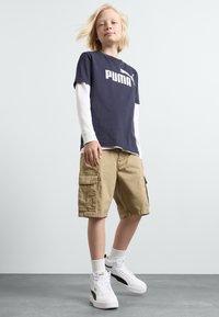Mornarsko plavi pamučni T-shirt s bijelim logotipom "PUMA", složen preko bijele majice dugih rukava. Bež cargo kratke hlače s bočnim džepovima. Bijele tenisice s crnim detaljima.