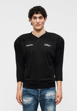 Νέος άνδρας με σγουρά μαλλιά που φορά μαύρο πουκάμισο με μακριά μανίκια με τα λογότυπα «DSQUARED2» και «ICONique», συνδυασμένο με φθαρμένα μπλε τζιν.