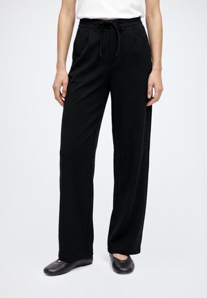 Femme portant un pantalon noir à jambes larges avec un nœud à l'avant et des chaussures plates noires, debout devant un fond clair uni.