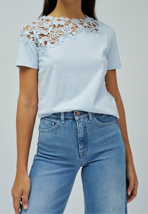 Femme portant un haut bleu clair à manches courtes avec des détails en dentelle florale sur l'épaule, associé à un jean bleu taille haute.