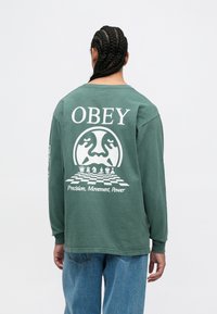 Garās piedurknes zaļa kokvilnas krekls ar baltu "OBEY" tekstu un šaha galda grafiku uz muguras, ar apaļu logo un vārdiem "Precizitāte, Kustība, Spēks."