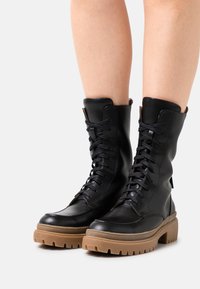 Stivali da combattimento in pelle nera con lacci e suola spessa marrone. Presentano un gambale alto e una texture liscia, con dettagli di cucitura sottili.