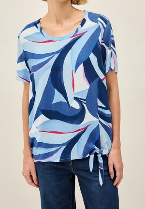 Frau trägt eine kurzärmelige, blau abstrakt gemusterte Bluse mit rundem Ausschnitt und seitlichem Knoten, kombiniert mit dunkelblauen Jeans.