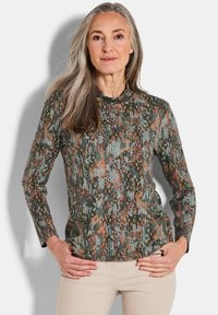 Vrouw met lang grijs haar die een gestreepte lange mouwen top en beige broek draagt, met handen op de heupen, tegen een effen achtergrond.