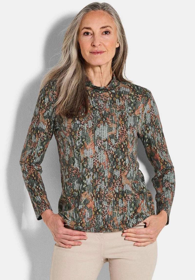 Vrouw met lang grijs haar die een gestreepte lange mouwen top en beige broek draagt, met handen op de heupen, tegen een effen achtergrond.