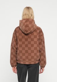 Persona bionda che indossa una giacca a quadri di velluto a coste marrone con cappuccio e pantaloni neri, di spalle contro uno sfondo bianco.