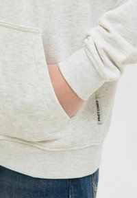 Helles graues Sweatshirt mit einer Fronttasche, ausgestattet mit gerippten Bündchen und einem Etikett an der Seite. Der Stoff hat eine weiche, strukturierte Oberfläche.