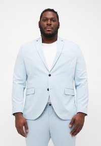 Helles blaues Anzug mit Slim Fit, Reverskragen, zwei Knöpfen und seitlichen Taschen. Kombiniert mit einem weißen T-Shirt darunter. Weiche Textur.
