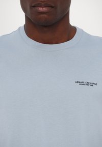 Gaiši zilas kokvilnas t-krekls ar apaļu kaklu un īsām piedurknēm. Uz krūts ir neliels melns drukāts logotips "ARMANI EXCHANGE".