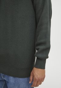 Pulover tricotat de culoare verde închis, cu o textură rib și mâneci lungi, având un tiv ajustat. Detalii vizibile de cusătură și un mic logo pe lateral.