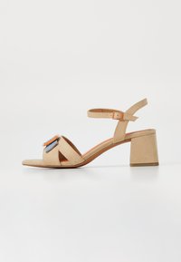 Sandalia de ante beige, destalonada y con un tacón grueso. Presenta una amplia correa en el tobillo y acentos decorativos en azul y naranja.