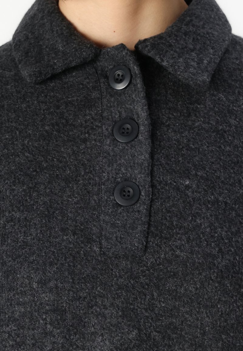 Haut en laine gris foncé avec col, présentant trois boutons noirs, tissu texturé avec une finition douce et un design de col structuré.